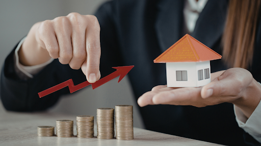 Person holding a small house model and upward red arrow above stacked coins, symbolizing rising home values or investment growth.